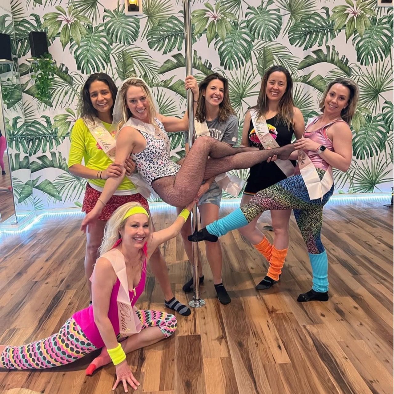 Six women in colorful 80s-style workout outfits and sashes posing around a dance pole in a bright studio with palm-leaf wallpaper and hardwood floors, playful bachelorette pole-dancing party vibe.