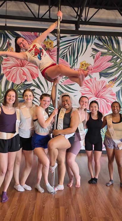 Nine women in activewear pose in a pole fitness studio with a bold pink tropical floral mural; one dancer holds a horizontal pole trick while the group smiles on a hardwood floor.