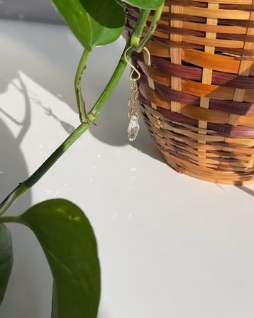 Sunlit wicker basket with a trailing pothos vine and a dangling gold-chain crystal plant charm casting tiny sparkles on a white tabletop