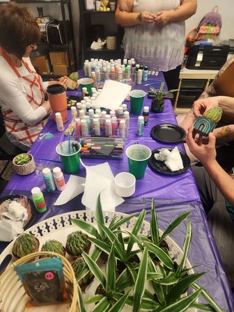 Indoor succulent-painting workshop with people decorating small cactus and decorative pots on a purple-covered table surrounded by acrylic paints, brushes, green cups and potted plants.