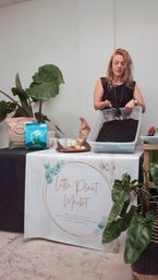 Indoor plant workshop: person scooping potting soil into a plastic tray at a market table surrounded by soil bags, tropical potted plants and potting tools.