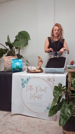 Indoor plant workshop: person scooping potting soil into a plastic tray at a market table surrounded by soil bags, tropical potted plants and potting tools.
