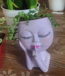 Lavender ceramic face planter in a shushing pose with painted eyelashes and pink lips, filled with a trailing round-leaf succulent, sitting on a wooden tabletop as indoor plant decor.