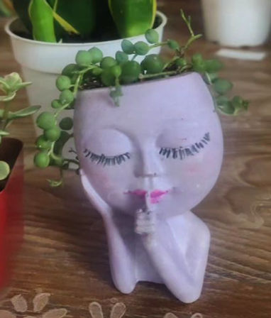 Lavender ceramic face planter in a shushing pose with painted eyelashes and pink lips, filled with a trailing round-leaf succulent, sitting on a wooden tabletop as indoor plant decor.