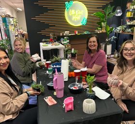 Smiling participants at an indoor plant workshop potting succulents and crafting DIY planters around a table with pots, tools, and a neon plant-themed sign.
