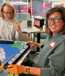 Two women playing mahjong at a casual cafe table — smiling woman with glasses arranges tiles on a colorful mat.