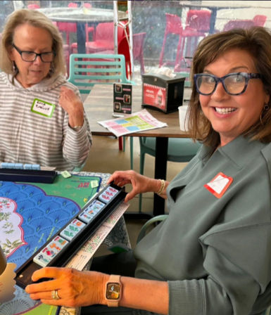 Two women playing mahjong at a casual cafe table — smiling woman with glasses arranges tiles on a colorful mat.
