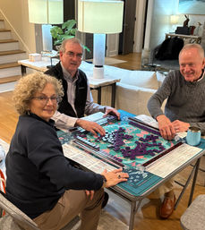 Three adults smiling and playing mahjong with purple tiles on a colorful game mat at a folding table in a cozy modern living room with lamps, sofa and staircase visible