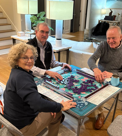 Three adults smiling and playing mahjong with purple tiles on a colorful game mat at a folding table in a cozy modern living room with lamps, sofa and staircase visible