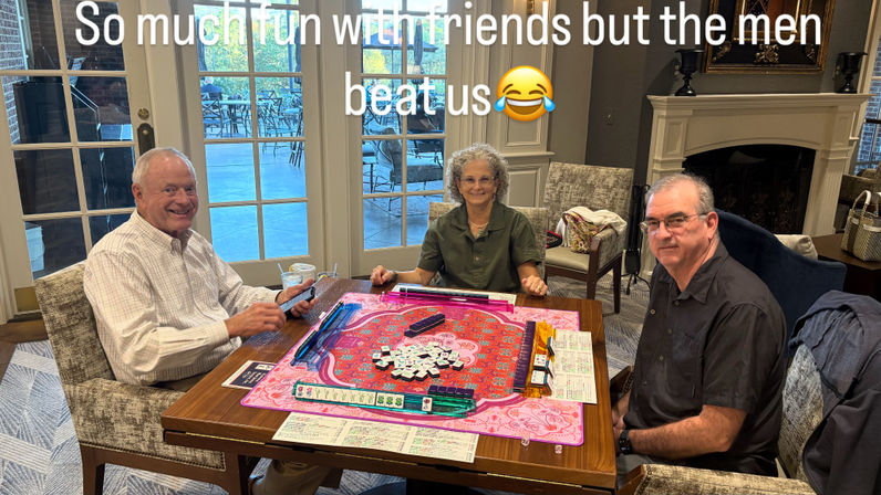 Three friends smiling around a table playing mahjong on a pink mat with tiles and racks, in a cozy clubhouse lounge with a fireplace and glass doors to a patio.