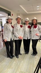 Four women wearing matching silver sequin jackets, white fedoras, black pants and red poinsettia corsages smiling together in a bright indoor event space — holiday performance look.