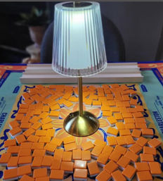 Brass table lamp casting a warm glow over a colorful tabletop game board covered in scattered orange rectangular tiles arranged like a playful mosaic.