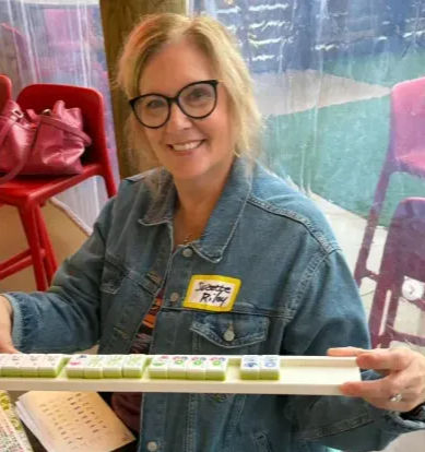 Smiling woman wearing glasses and a denim jacket holding a rack of mahjong tiles inside a colorful cafe patio with red chairs and a pink bag nearby