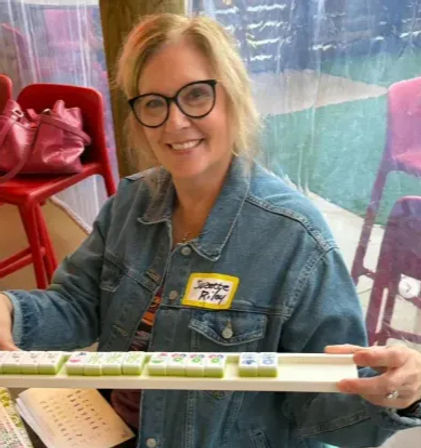 Smiling woman wearing glasses and a denim jacket holding a rack of mahjong tiles inside a colorful cafe patio with red chairs and a pink bag nearby