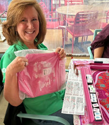 Smiling woman in a green shirt seated at an outdoor patio table holding a sealed pink gift bag with white script; pink tile game set, score sheets and glasses on the table and red patio chairs in the background.