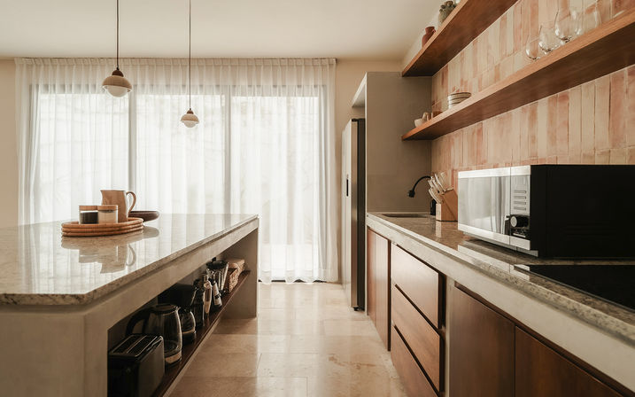 Sunlit modern kitchen with a polished marble island, hanging pendant lights, warm wooden cabinets, textured tile backsplash and open shelves with glassware, microwave and small appliances, sheer floor-to-ceiling curtains.