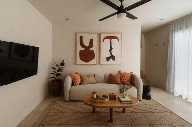 Cozy modern neutral living room with curved beige sofa, rust-orange throw pillows, round wooden coffee table with decorative tray and books, abstract brown wall art, potted plant, ceiling fan and sheer curtains filtering daylight