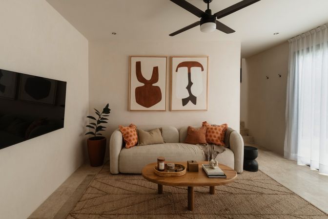 Cozy modern neutral living room with curved beige sofa, rust-orange throw pillows, round wooden coffee table with decorative tray and books, abstract brown wall art, potted plant, ceiling fan and sheer curtains filtering daylight