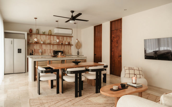 Sunlit open-plan kitchen and dining area with natural wood table and black-accent chairs, marble island and tiled backsplash, pendant lights, ceiling fan, neutral beige tones and a cozy living nook with wall-mounted TV.