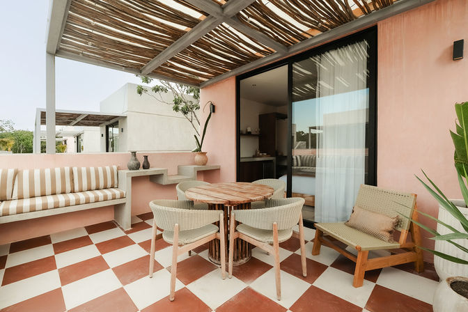 Sunny boho outdoor terrace with pink stucco wall, wooden pergola, red-and-white checkerboard floor, round wooden dining table with woven chairs and striped bench