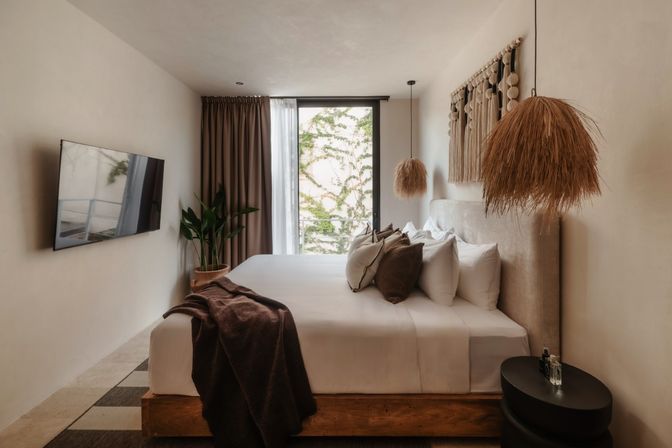 Modern boho bedroom with king bed and layered pillows, neutral linens, woven pendant lights, potted plant, wall-mounted TV and floor-to-ceiling window framed by ivy