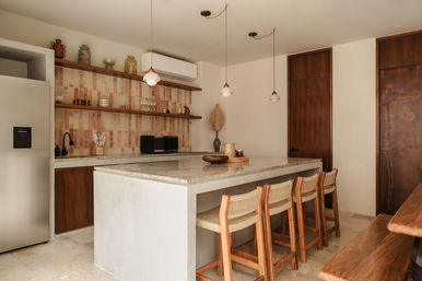 Warm minimalist kitchen with large stone island and four wooden woven bar stools, hanging pendant lights, terracotta tiled backsplash, open wooden shelves with pottery, stainless fridge and microwave.
