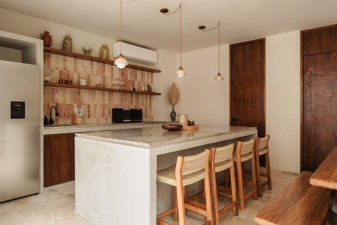 Warm minimalist kitchen with large stone island and four wooden woven bar stools, hanging pendant lights, terracotta tiled backsplash, open wooden shelves with pottery, stainless fridge and microwave.