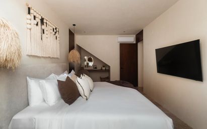 Cozy modern neutral bedroom with king bed in white linens, brown accent pillows, macramé wall hanging, straw pendant lamps, small desk under a sloped nook, and wall-mounted flat-screen TV