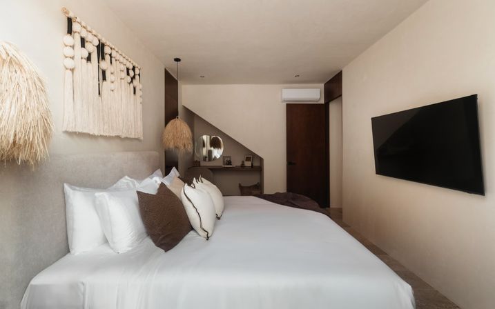 Cozy modern neutral bedroom with king bed in white linens, brown accent pillows, macramé wall hanging, straw pendant lamps, small desk under a sloped nook, and wall-mounted flat-screen TV