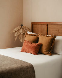 Cozy boho bedroom corner with a rattan headboard, hanging woven palm pendant light and warm terracotta and rust textured pillows on crisp white bedding.