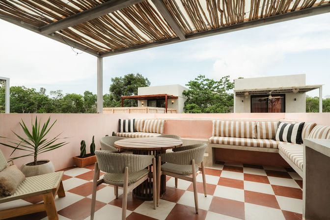 Rooftop terrace with woven pergola, round wooden dining table and wicker chairs, built-in bench with striped cushions, red-and-white checkerboard tile floor, potted succulents and leafy green trees beyond.