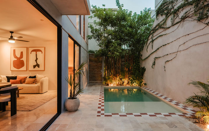 Cozy modern backyard courtyard at dusk with sliding glass doors to a warm-lit living room, tiled patio and narrow plunge pool edged in red-and-white tile, potted palms, lush plants and vine-covered wall.