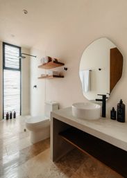 Sunlit modern minimalist bathroom with concrete vanity and round white vessel sink, matte-black faucet, organic-shaped mirror, floating wooden shelves with rolled towels, walk-in rainfall shower and textured frosted window over warm stone tile floor.