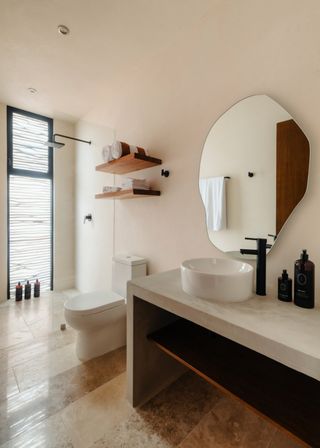 Sunlit modern minimalist bathroom with concrete vanity and round white vessel sink, matte-black faucet, organic-shaped mirror, floating wooden shelves with rolled towels, walk-in rainfall shower and textured frosted window over warm stone tile floor.