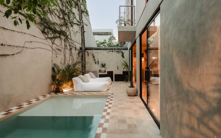Modern urban courtyard with a small plunge pool and tiled patio, white cushioned loungers, potted palms and vine-covered walls beside warm-lit sliding glass doors.