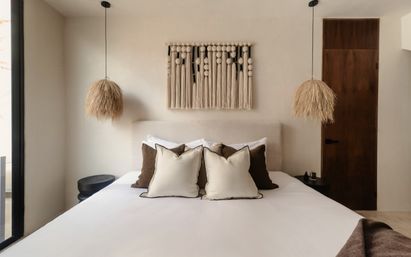 Cozy minimalist boho bedroom with a neutral upholstered bed, layered cream and brown pillows, large macramé wall hanging, and two straw pendant lights flanking the bed