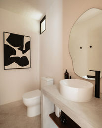 Minimalist modern powder room with white vessel sink, matte black faucet, organic-shaped mirror, black-and-white abstract artwork and compact toilet on warm neutral walls.