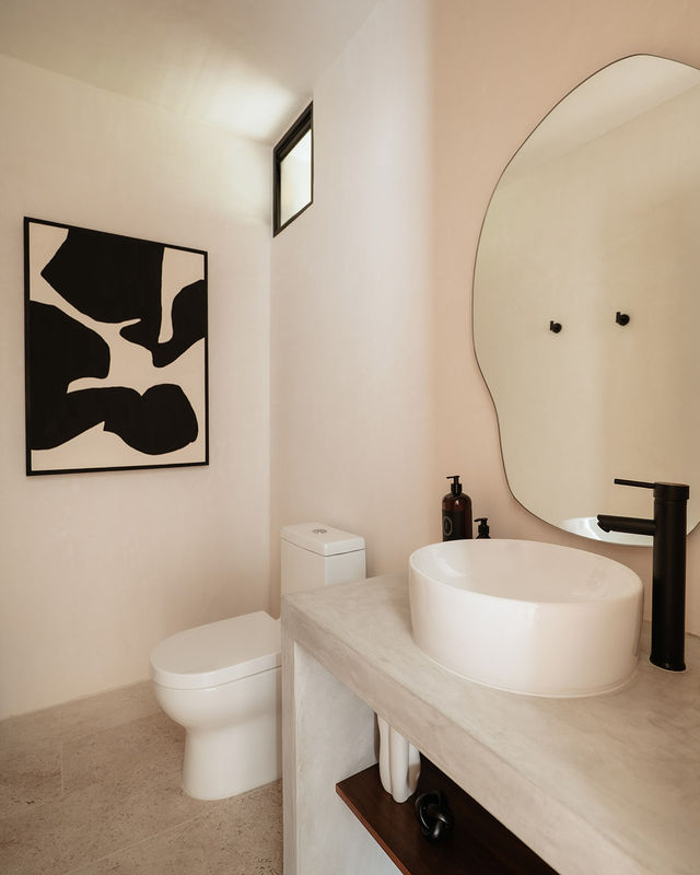 Minimalist modern powder room with white vessel sink, matte black faucet, organic-shaped mirror, black-and-white abstract artwork and compact toilet on warm neutral walls.
