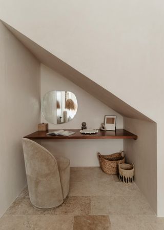 Cozy minimalist home office alcove under a slanted ceiling with a floating wooden desk, round mirror, beige upholstered chair, open book and woven baskets in neutral boho tones