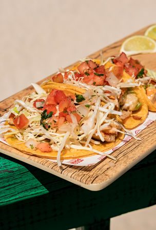 Three Mexican-style grilled fish tacos on double corn tortillas topped with shredded cabbage and fresh pico de gallo, served on a wooden board with lime wedges — bright coastal street-food styling
