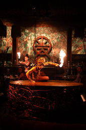 Fire dancer in tribal costume crouches on a round vine-wrapped stage table in a dim tiki-style bar, performing with a flaming torch while patrons watch under warm orange lighting.