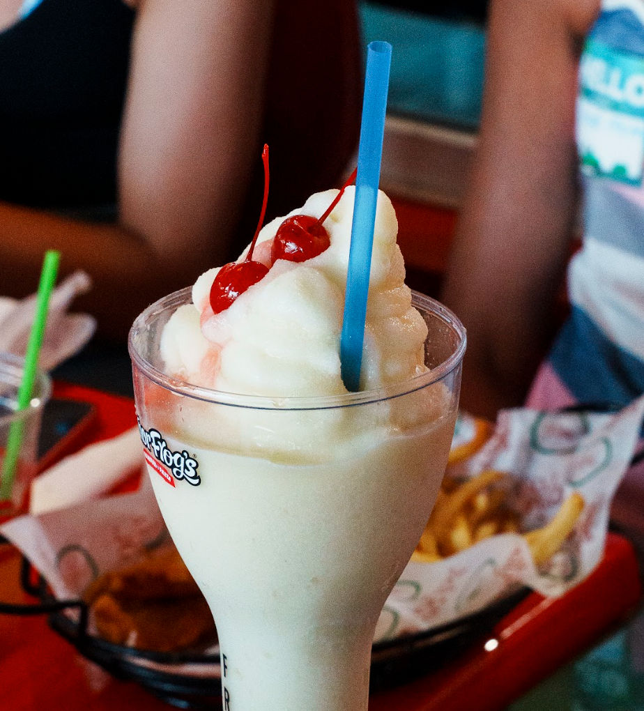 Creamy frozen vanilla milkshake in a tall clear glass topped with two maraschino cherries and a blue straw, served on a casual diner table with fries in the background.