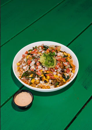 Colorful Mexican-style rice bowl with grilled corn, sautéed peppers and onions, pico de gallo, crumbled cheese and a scoop of guacamole, served with a small cup of creamy sauce on a bright green table.