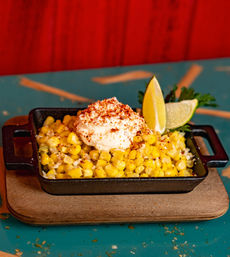 Cast-iron skillet of elote-style Mexican street corn — buttery kernels topped with creamy cheese, chili powder, lime wedges and parsley on a wooden board
