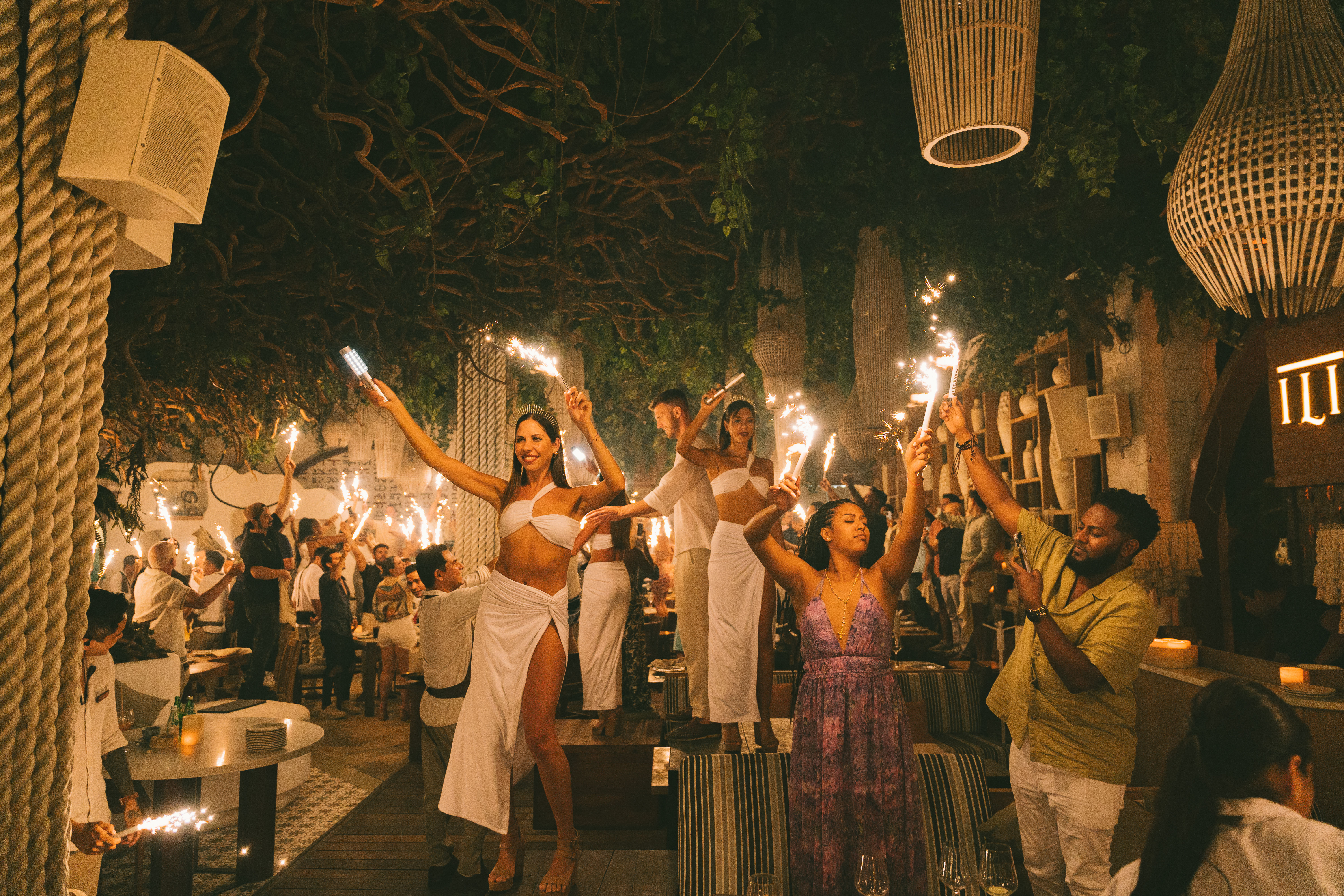 Nighttime beach-club restaurant scene with dancers in white standing on tables waving sparklers under a leafy canopy and wicker lamps, guests cheering and taking photos.