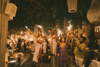 Nighttime beach-club restaurant scene with dancers in white standing on tables waving sparklers under a leafy canopy and wicker lamps, guests cheering and taking photos.