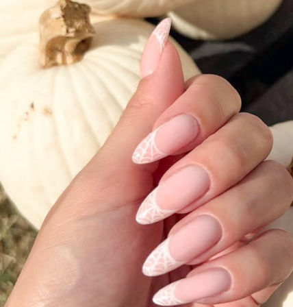 Matte blush stiletto nails with delicate white spiderweb tip art, a chic fall/Halloween manicure posed against a white pumpkin backdrop.