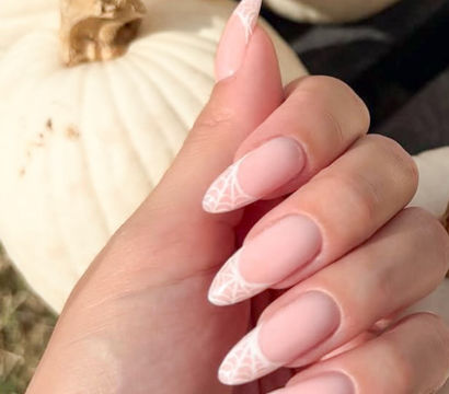 Matte blush stiletto nails with delicate white spiderweb tip art, a chic fall/Halloween manicure posed against a white pumpkin backdrop.