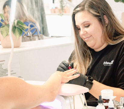 Manicurist painting pink nails under an LED curing lamp in a bright, plant‑accented nail salon with manicure bottles on the table.