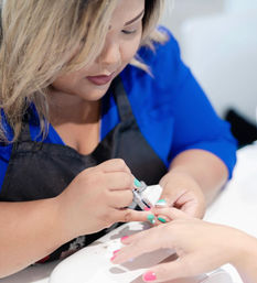 Close-up of a nail technician in a blue top painting a client's bright pink and mint manicure over a nail dryer in a salon